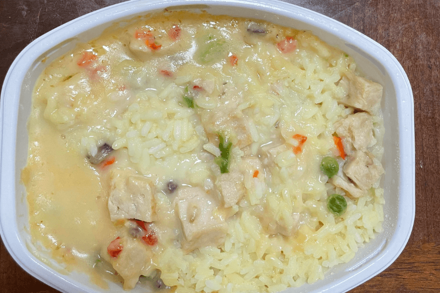 A microwaved frozen meal in a white plastic tray, featuring creamy chicken and rice. The dish contains chunks of white chicken, yellow rice, peas, carrots, green beans, and a thick, beige gravy-like sauce.