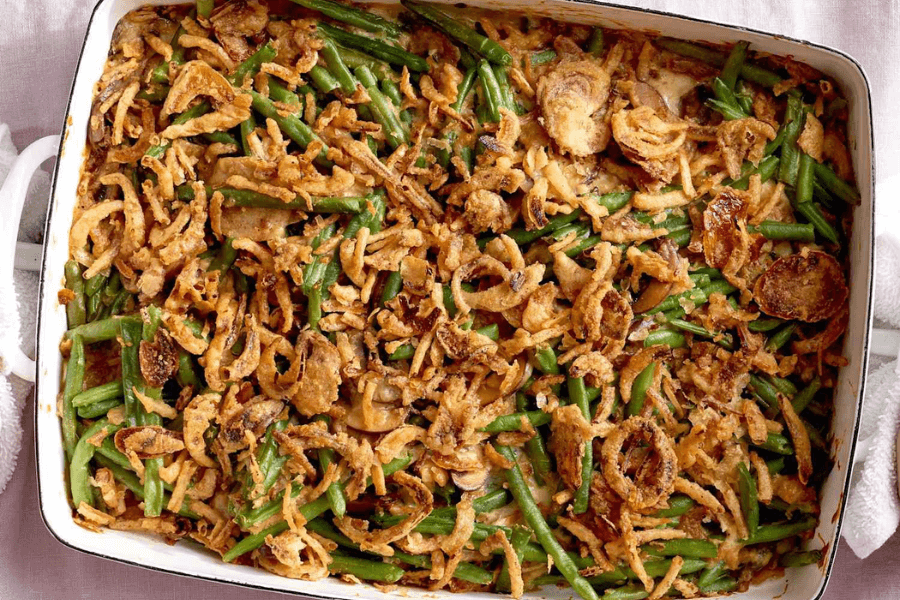 A rectangular white baking dish filled with green bean casserole, featuring vibrant green beans mixed with creamy sauce and topped with a generous layer of crispy fried onions. The golden-brown topping is slightly charred in spots, adding texture and crunch.