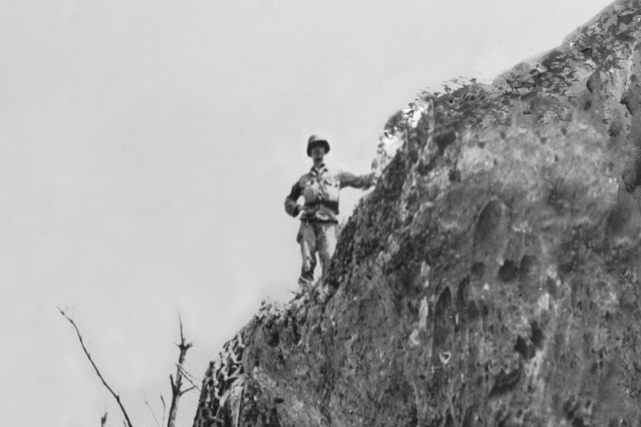 Black-and-white photo of a World War II soldier standing near the edge of a steep cliff, gripping the rocky surface for support. The image captures a dramatic and elevated vantage point with barren branches visible in the lower left and an expansive, blank sky overhead.