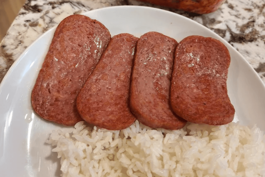 A white plate with four slices of pan-fried Spam placed next to a serving of steamed white rice. The Spam is golden brown with a slight crisp on the edges, and the rice appears fluffy and slightly sticky.