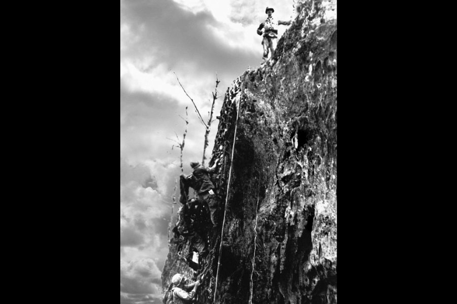 Dramatic black-and-white photo of World War II soldiers scaling a steep, jagged cliff using a rope, while another soldier stands watchfully at the top. The rugged terrain and cloudy sky emphasize the intensity and danger of the climb, likely referencing the infamous ascent at Hacksaw Ridge.