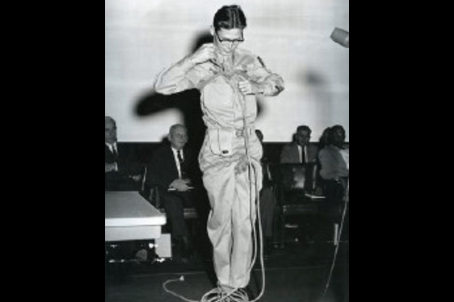 Black-and-white photo of a man in a military-style uniform standing on a stage, demonstrating how to tie a rope harness. He appears focused and methodical, with several seated individuals watching him from behind, suggesting a public or educational event.