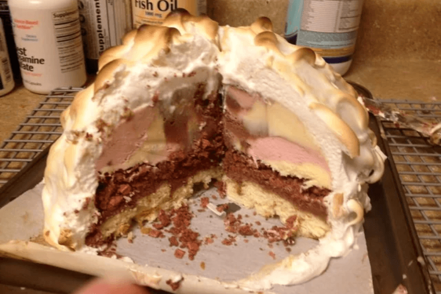 A partially sliced Baked Alaska revealing layers of yellow cake, chocolate cake, multiple flavors of ice cream including vanilla, chocolate, and strawberry, all encased in a toasted meringue shell. The dessert sits on a baking sheet with crumbs and melted ice cream around the base.