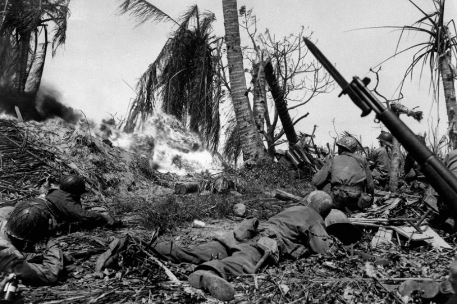 Intense black-and-white combat photo of U.S. soldiers on the ground during a World War II Pacific battle, taking cover as flames erupt on a palm-covered hill. Fallen soldiers, rifles with bayonets, and debris are scattered in the foreground, while smoke and fire dominate the chaotic jungle backdrop.