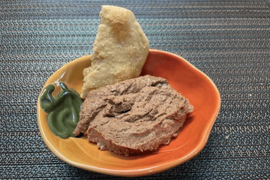 A small orange dish with a scoop of liver pâté, a piece of crusty bread or cracker, and a swirl of green wasabi paste. The spread has a rich, smooth texture with visible chunks, served on a woven placemat.