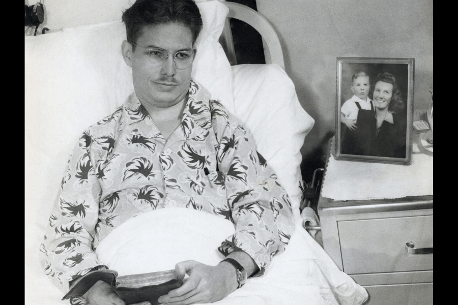 Black-and-white photo of a man resting in a hospital bed, wearing a patterned pajama top and holding a Bible in his lap. Beside him on a nightstand is a framed photo of a smiling woman holding a young child, adding a personal and emotional touch to the hospital setting.