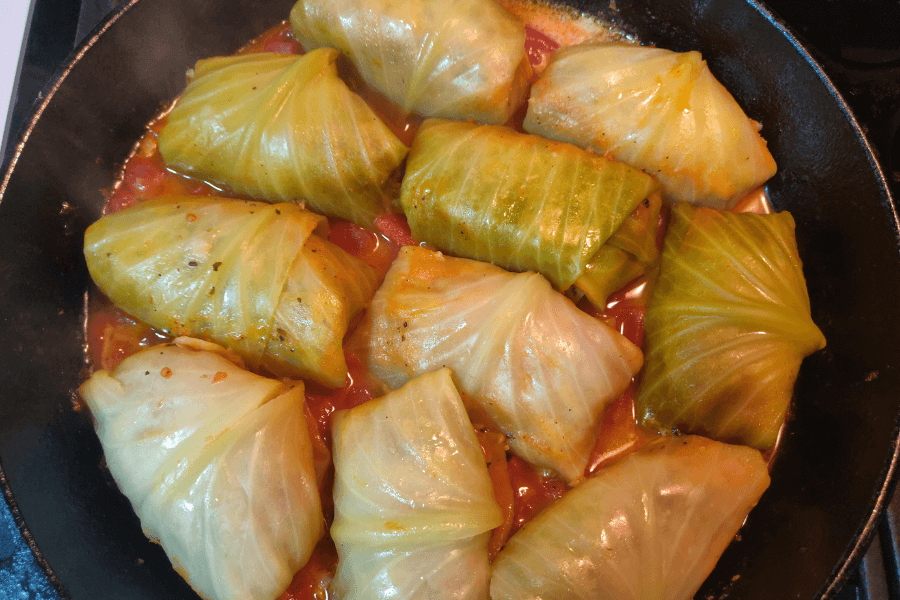 A skillet filled with cabbage rolls simmering in a tomato-based sauce. Each roll is tightly wrapped in a steamed cabbage leaf, with visible seams and a slight golden tint from cooking.