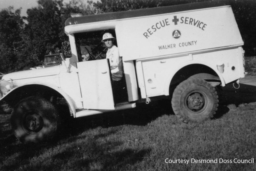 Black-and-white photo of a vintage rescue vehicle labeled “Rescue Service Walker County” with a red cross and civil defense insignia. A uniformed individual wearing a helmet sits in the driver’s seat with the door open, facing the camera. Text in the bottom corner reads “Courtesy Desmond Doss Council.”
