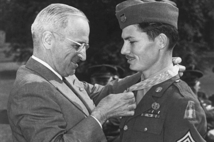Black-and-white photo of President Harry S. Truman smiling as he places the Medal of Honor around the neck of Army Corporal Desmond Doss, who is dressed in full uniform with multiple service ribbons and patches. The outdoor ceremony captures a historic moment of recognition for Doss’s heroic acts as a combat medic during World War II.