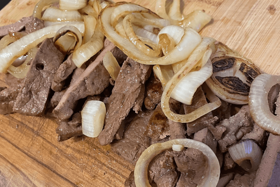 Sliced beef liver with sautéed onions served on a wooden cutting board. The liver is cooked medium and topped with both tender and slightly charred onion slices.