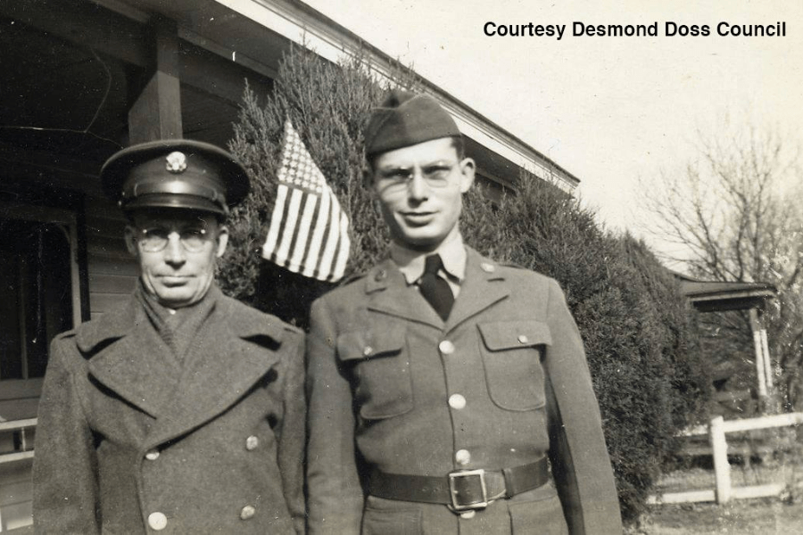 Black-and-white photo of two uniformed men standing outside a house, one wearing a garrison cap and the other a round military cap. An American flag is displayed in the background near some large bushes, and both men wear glasses and military coats. Text in the corner reads, "Courtesy Desmond Doss Council."