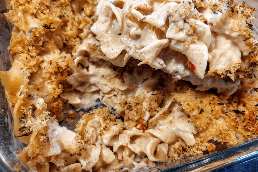 A creamy baked noodle casserole in a glass dish, topped with a golden brown breadcrumb crust. The dish contains wide egg noodles mixed with a thick white sauce, herbs, and bits of vegetables or meat, partially scooped out to reveal the creamy interior.