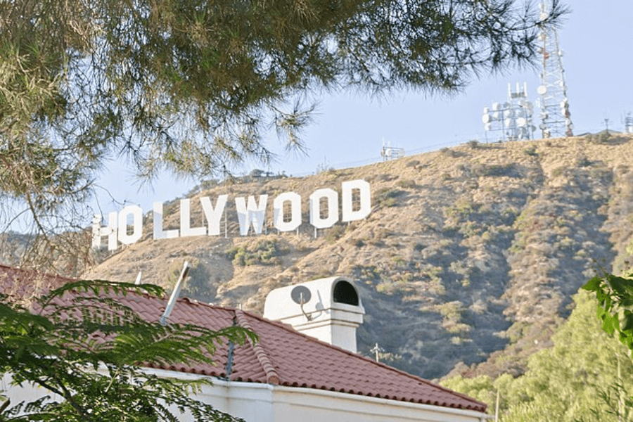 Daytime photo of the iconic white Hollywood Sign perched on the hillside in Los Angeles, California, partially framed by pine tree branches in the foreground. A red-tiled rooftop and chimney with satellite equipment are visible at the bottom of the image, while communication towers sit atop the hill.