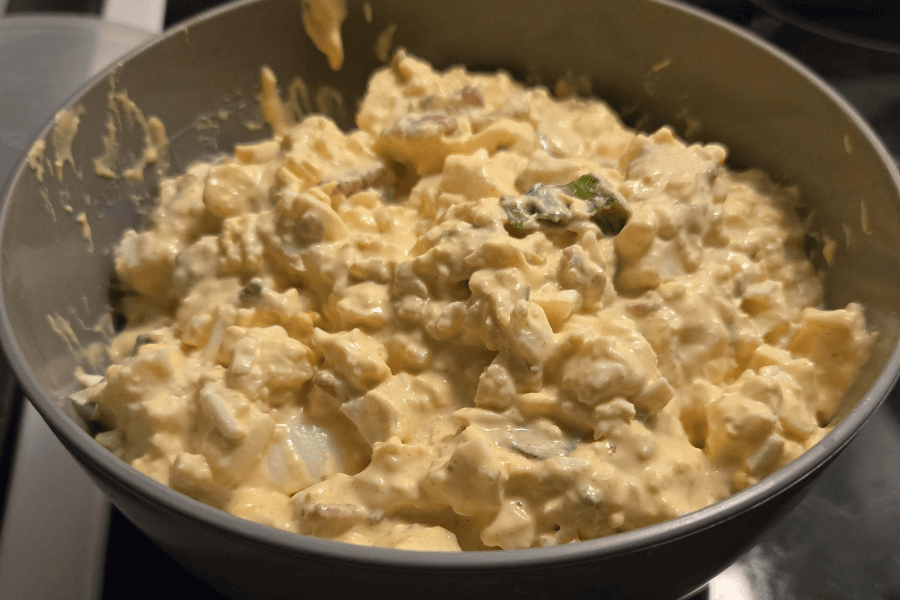 A large bowl filled with chunky homemade potato salad, featuring diced potatoes, hard-boiled eggs, chopped pickles, red onion, and green herbs, all coated in a creamy yellow mayonnaise-based dressing.