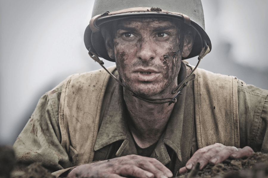 Close-up of a battle-worn soldier with dirt and blood on his face, wearing a World War II-era helmet and uniform. His expression is tense and focused as he crouches in a war zone, gripping the earth with both hands.