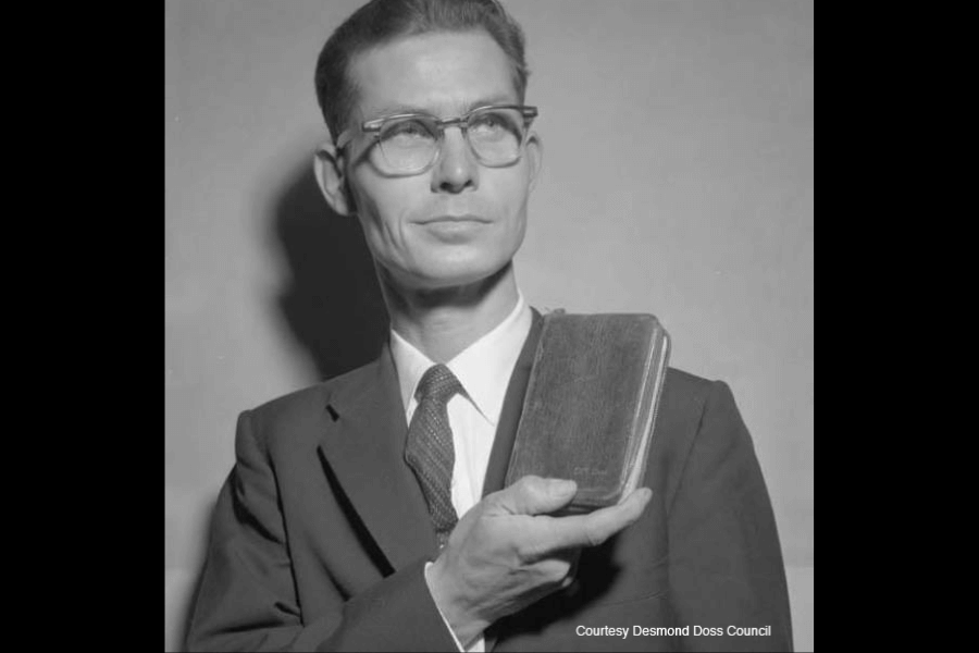 Black-and-white portrait of Desmond Doss in a suit and tie, holding a Bible upright against his chest with a composed, steadfast expression. He wears glasses and is lit from the side, casting a strong shadow behind him. Text at the bottom reads, “Courtesy Desmond Doss Council.”