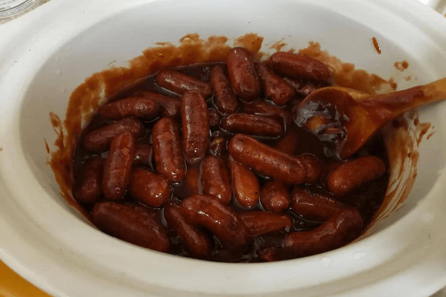 A white slow cooker filled with cocktail sausages simmering in a thick, glossy brown barbecue sauce. A wooden spoon coated with sauce rests inside the crock, suggesting the dish is freshly stirred and still warm.