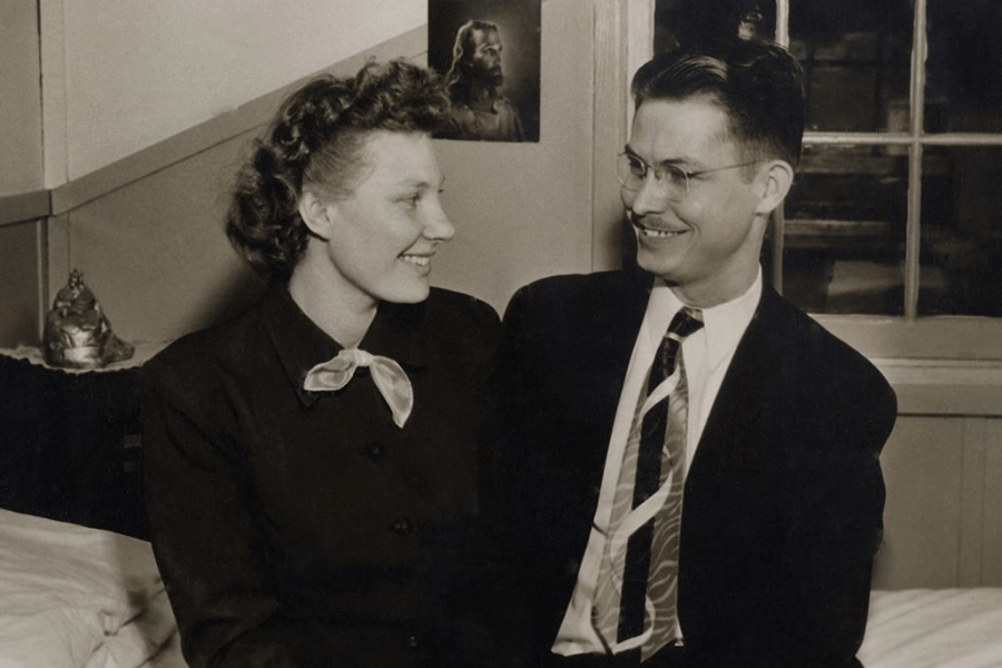 Sepia-toned photo of a smiling couple sitting close together on a bed, gazing at each other warmly. The woman wears a dark dress with a light bow at the collar, and the man wears glasses, a suit, and a striped tie. Behind them, a picture of Jesus hangs on the wall near a window.