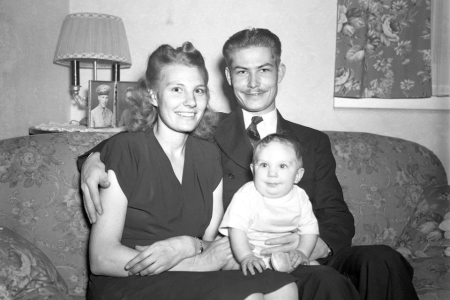 Black-and-white family photo of Desmond Doss sitting on a floral couch with his wife Dorothy and their baby boy. Dorothy wears a short-sleeved dress, Desmond is in a suit and tie, and the baby is smiling in a white shirt. A framed photo of a soldier and a table lamp are visible in the background beside floral curtains.