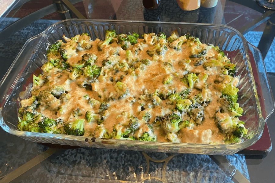 A glass baking dish filled with broccoli casserole, featuring bright green broccoli florets coated in a creamy cheese sauce and topped with melted, slightly golden cheese. The dish is placed on a glass-top table.