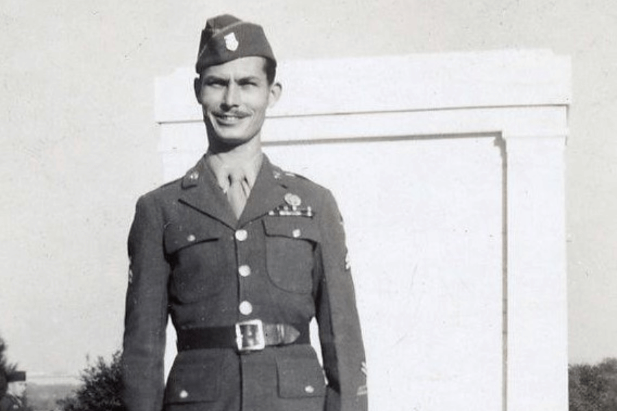Vintage black-and-white photo of Desmond Doss in full U.S. Army dress uniform, standing outdoors in front of a white monument or structure. He wears a garrison cap, decorated with medals and service ribbons, and smiles confidently at the camera.