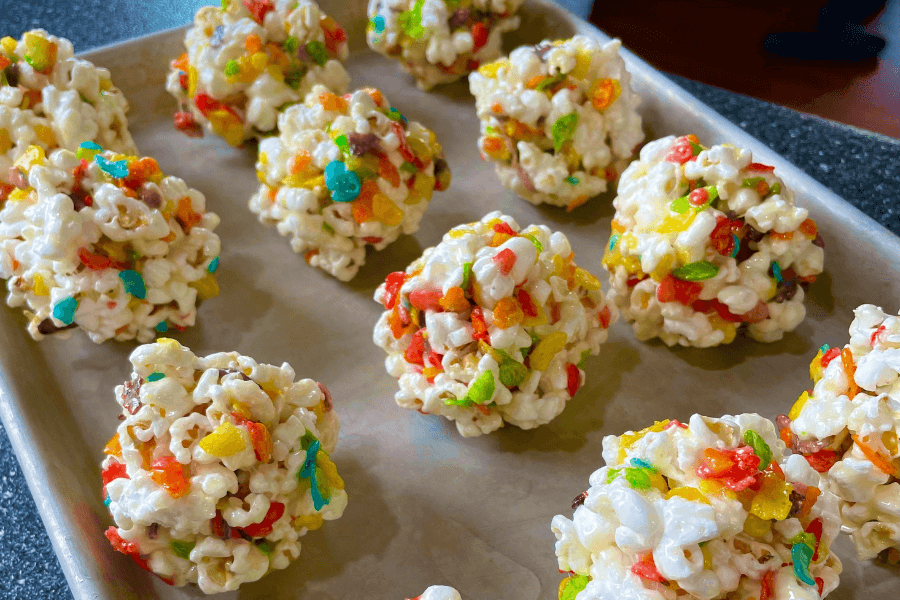 A tray of colorful popcorn balls made with melted marshmallows and Fruity Pebbles cereal. Each treat is shaped into a round cluster and set on parchment paper for cooling.