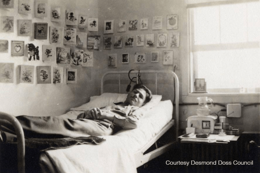 Black-and-white photo of Desmond Doss lying in a hospital bed, arms folded across his chest, in a sunlit room decorated with dozens of greeting cards covering the wall above him. A window on the right lets in natural light, and a small nightstand holds a water pitcher, radio, and personal items. Text in the corner reads, “Courtesy Desmond Doss Council.”