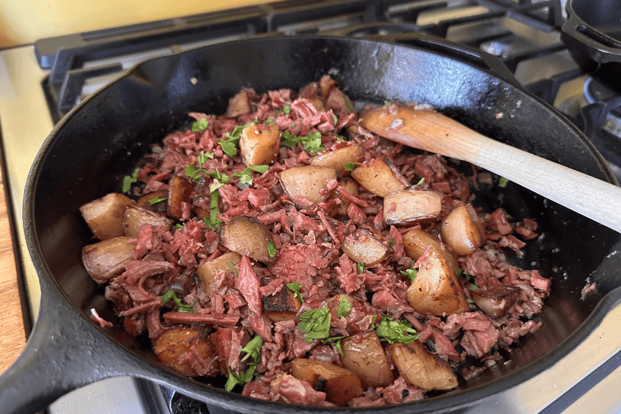 A cast iron skillet filled with corned beef hash, made with shredded corned beef, golden-brown cubed potatoes, and garnished with chopped fresh herbs. A wooden spoon rests in the skillet, suggesting the dish is freshly prepared and ready to serve.