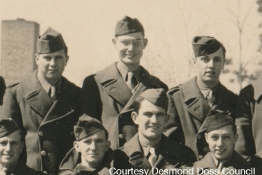 Sepia-toned group photo of young World War II soldiers dressed in double-breasted military overcoats and garrison caps, standing and crouching outdoors for a portrait. The background shows bare trees and a tall building, suggesting the image was taken during training or deployment preparation. Text reads, “Courtesy Desmond Doss Council.”