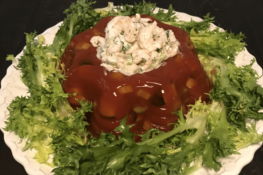 A retro-style savory gelatin mold made with tomato-based gelatin and embedded vegetables, surrounded by leafy green lettuce on a white platter. The center of the mold is filled with a creamy shrimp salad made with small shrimp, celery, and herbs.