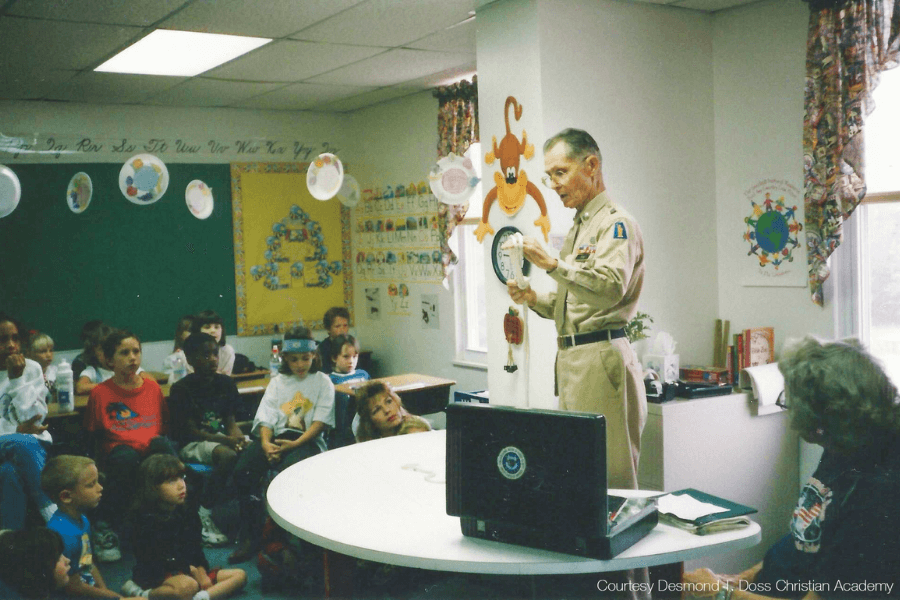 Color photo of Desmond Doss standing in uniform inside a brightly decorated classroom, speaking to a group of young children seated on the floor and at desks. He gestures toward a cartoon monkey wall clock, engaging the attentive students. A laptop and open binder sit on a round table in the foreground. Text in the corner reads, “Courtesy Desmond T. Doss Christian Academy.”