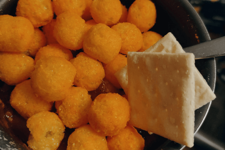 A bowl of chili topped with bright orange cheese balls, served with two saltine crackers resting on the edge of the bowl. The contrasting textures and colors give the dish a playful, unconventional presentation.