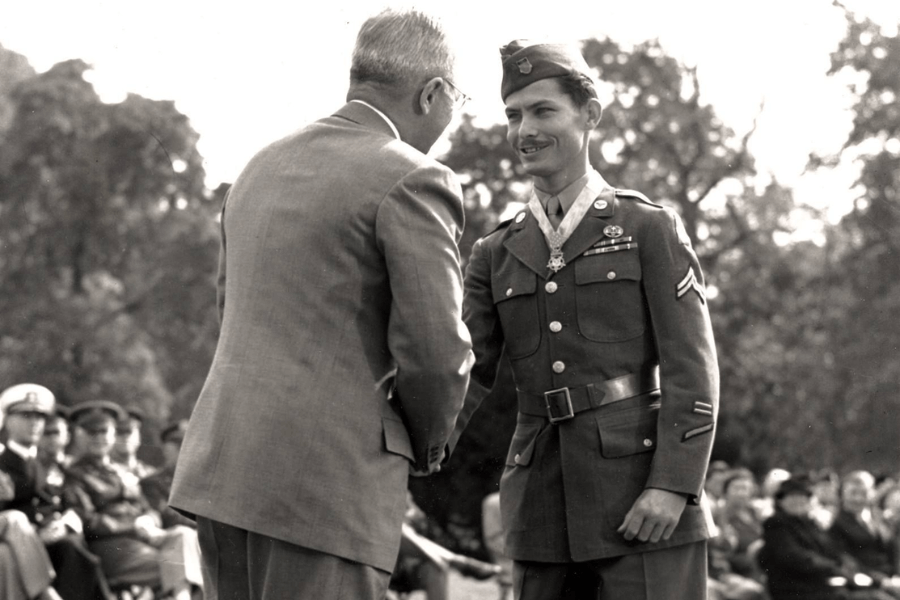 Black-and-white photo of President Harry S. Truman shaking hands with Corporal Desmond Doss during a formal Medal of Honor ceremony. Doss stands in full Army dress uniform, wearing the Medal of Honor around his neck and smiling, while an audience of military personnel and civilians watches in the background.