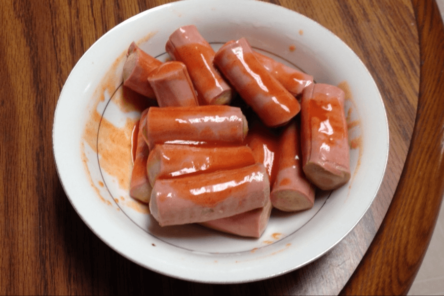 A white bowl filled with sliced hot dogs coated in a thin, reddish-orange sauce. The hot dogs are cut into short segments and appear to be served at room temperature on a wooden table.