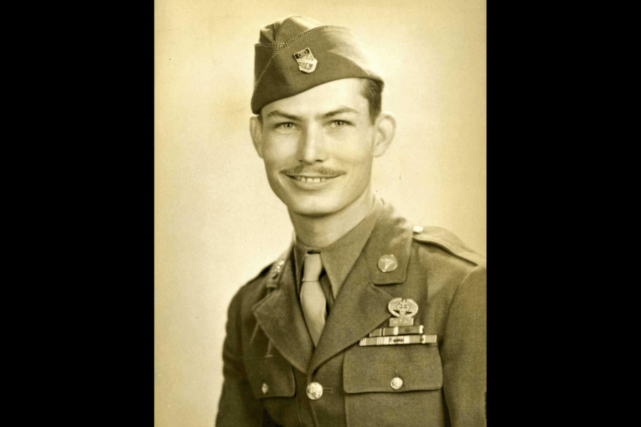 Sepia-toned portrait of Desmond Doss in his U.S. Army dress uniform, smiling slightly and looking directly at the camera. He wears a garrison cap, military ribbons, the Combat Medic Badge, and the Medal of Honor around his neck, symbolizing his heroic WWII service.
