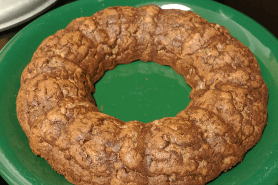 A dense, ring-shaped fruitcake with a rough, textured surface, sitting on a green plate. The cake appears to be homemade, with visible chunks of nuts and dried fruits baked into the golden-brown crust.
