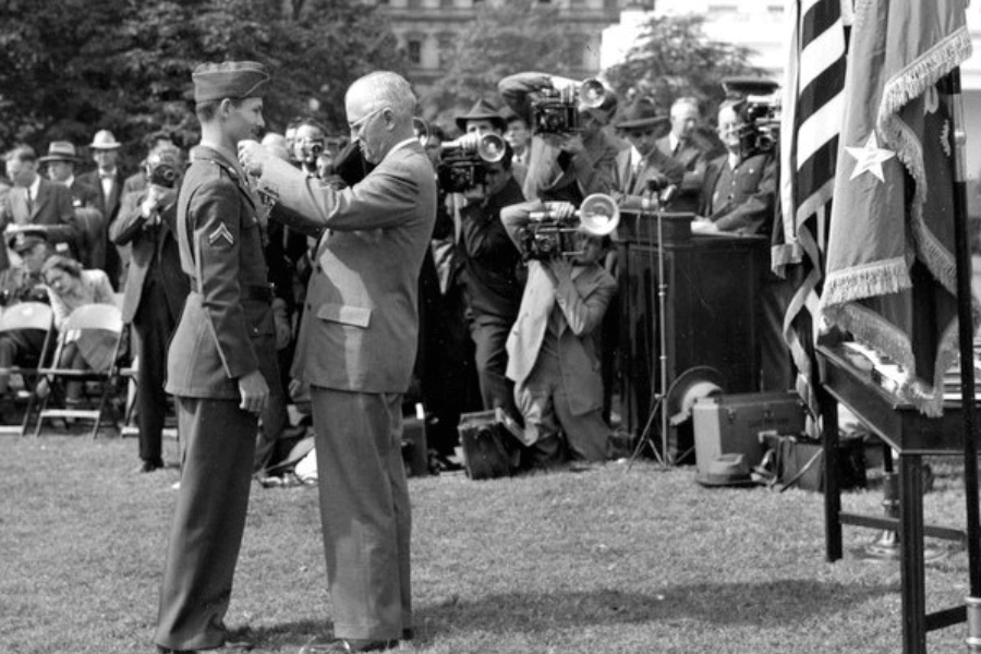 Historic black-and-white photo of President Harry S. Truman presenting the Medal of Honor to Corporal Desmond Doss in a formal outdoor ceremony. Doss stands in full uniform as photographers and reporters capture the moment, with American flags and military personnel visible in the background.