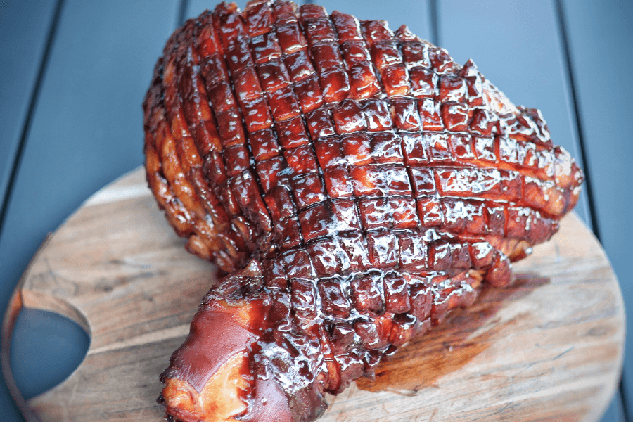 A glazed holiday ham with a beautifully caramelized exterior and a crosshatch pattern scored into the surface. The ham sits on a round wooden cutting board, showcasing its rich, glossy finish and crispy edges.