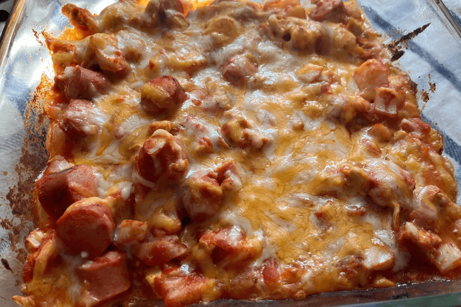 A cheesy hot dog casserole baked in a glass dish, filled with sliced hot dogs, tomato sauce, and melted cheese on top. The cheese is golden and bubbly, with slightly crispy edges where it has browned in the oven.