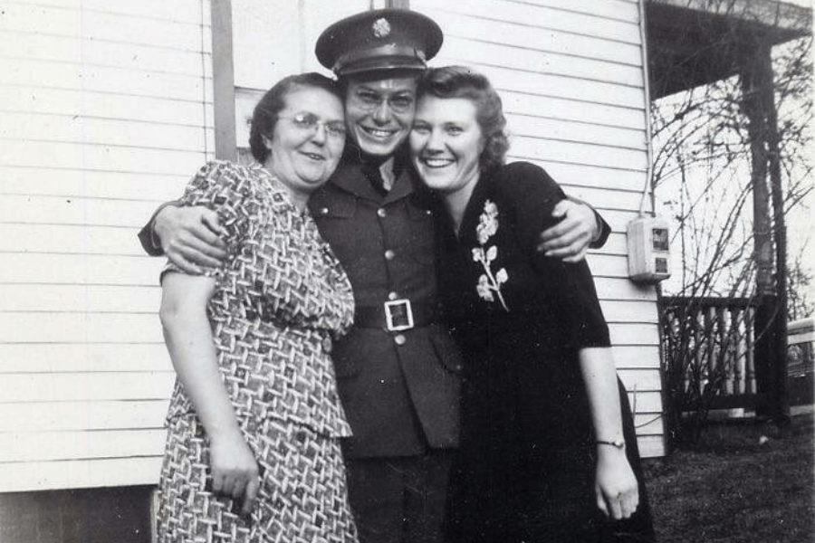Black-and-white photo of a smiling soldier in uniform standing between two women, wrapping his arms around them in a joyful embrace. The group poses in front of a white clapboard house, suggesting a heartfelt reunion at home during or after WWII.