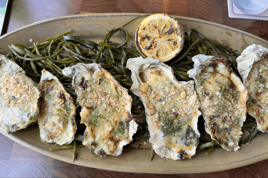 A platter of baked oysters served on a bed of seaweed, topped with a golden brown cheesy breadcrumb crust and garnished with fresh herbs. A grilled half-lemon sits at the center of the oval dish, ready for squeezing.
