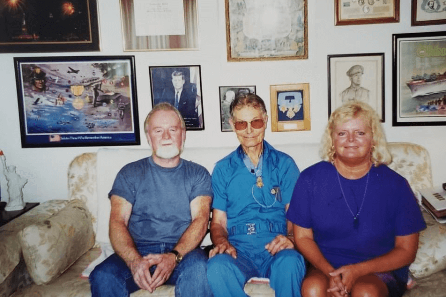Color photo of Desmond Doss seated between a man and a woman on a floral couch, wearing a blue outfit and his Medal of Honor around his neck. The background wall is filled with framed memorabilia, including military portraits, patriotic artwork, and historical photographs, highlighting his decorated legacy.
