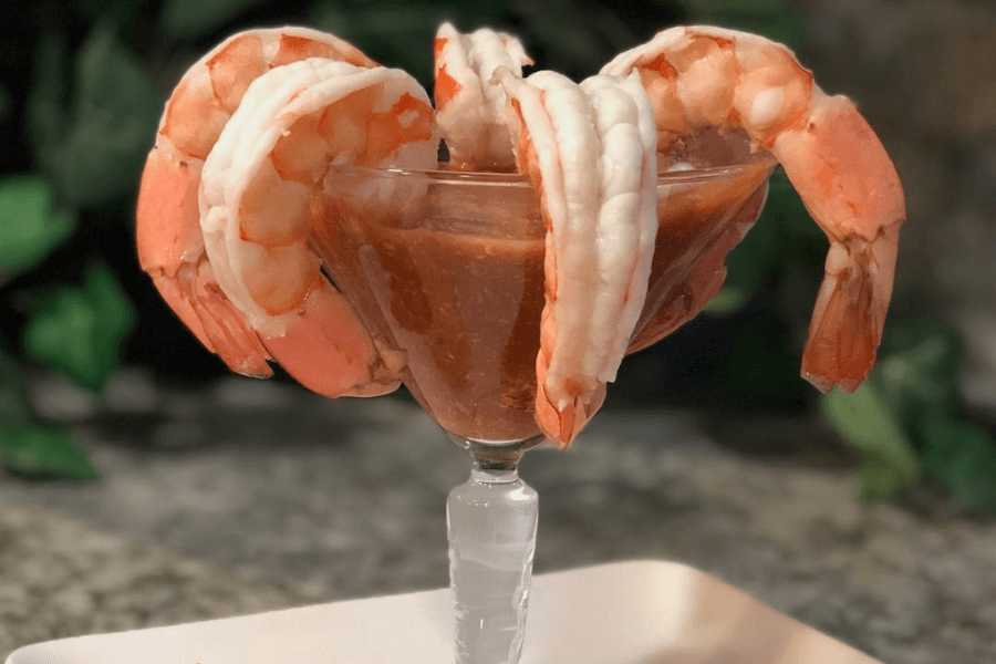 A classic shrimp cocktail served in a glass with a deep red cocktail sauce. Large, peeled shrimp are draped over the rim of the glass, ready to be dipped and eaten.