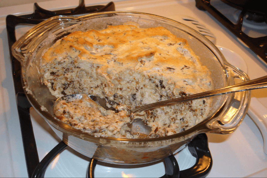 A glass baking dish filled with a baked wild rice casserole that has been partially scooped out, revealing a creamy mixture of rice, mushrooms, and possibly meat or nuts. The top is lightly browned and slightly crusty from baking.
