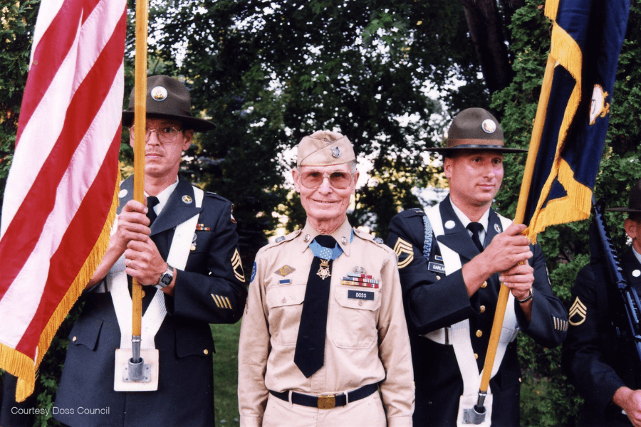 Color photograph of Desmond Doss, wearing a tan military uniform with his Medal of Honor and nameplate, standing between two uniformed servicemen holding the American flag and a unit banner. The group is part of a ceremonial color guard, posed outdoors in front of trees. The text “Courtesy Doss Council” appears in the bottom left corner.