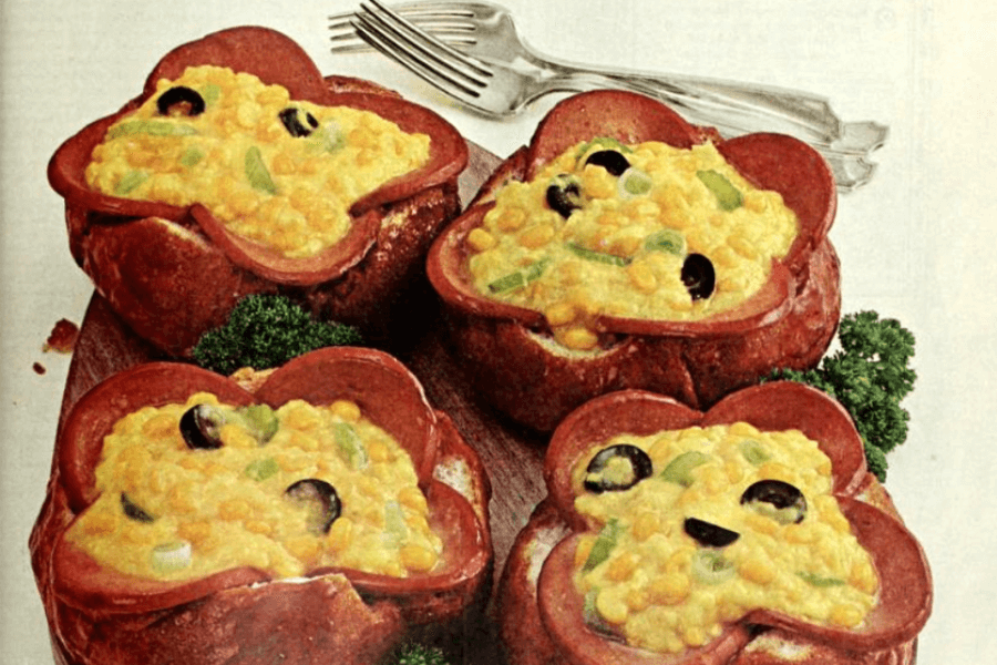 Four bread rolls hollowed out and filled with a creamy corn and cheese mixture, topped with sliced black olives and green bell peppers. Each bread cup is lined with slices of bologna arranged like flower petals, and the dish is garnished with parsley.