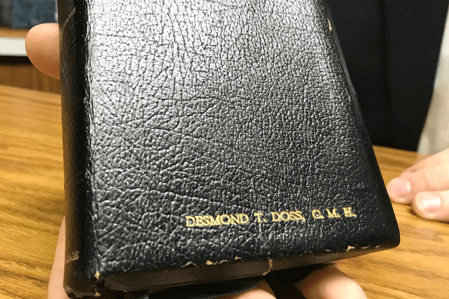 Close-up image of a black leather Bible with gold-stamped text reading “DESMOND T. DOSS C.M.H.” on the bottom corner of the cover. The Bible is being held gently over a wooden table by two hands, likely during a display or interview.
