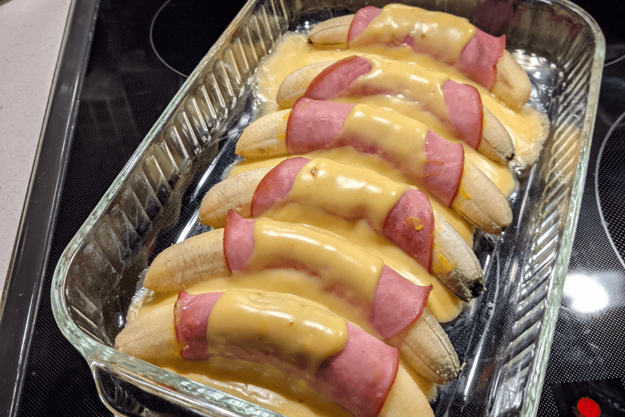 A glass baking dish containing whole bananas wrapped in slices of ham and topped with melted cheese sauce. The dish is arranged in neat rows and baked until the cheese is smooth and slightly browned at the edges.