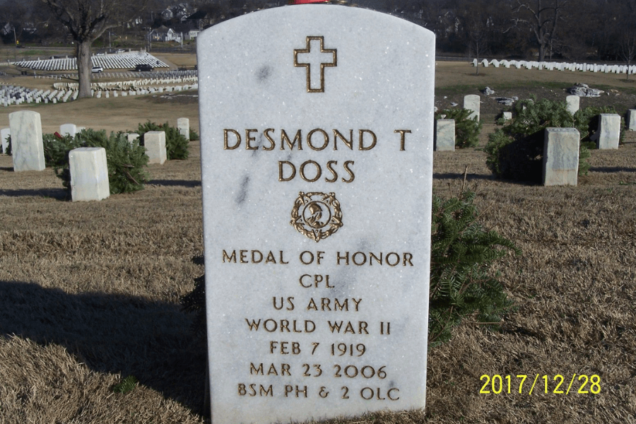 Gravestone of Desmond T. Doss in a military cemetery, engraved with a cross and inscribed with “Medal of Honor,” his rank (CPL), branch (US Army), and service in World War II. Dates of birth and death are February 7, 1919, and March 23, 2006, and it includes the initials for additional honors: BSM, PH & 2 OLC. Rows of similar headstones and wreaths are visible in the background.