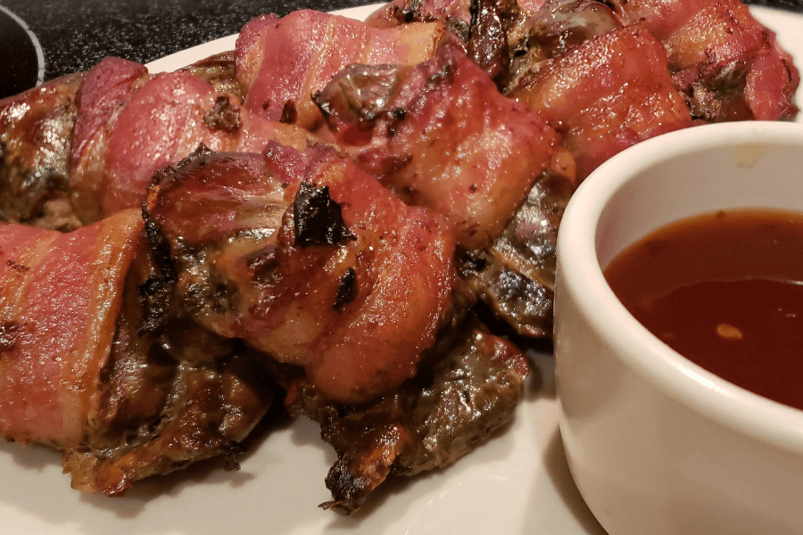 A plate of bacon-wrapped chicken livers, cooked to a deep golden brown with crispy edges. Served alongside a small white ramekin filled with dark dipping sauce, likely barbecue or sweet glaze.